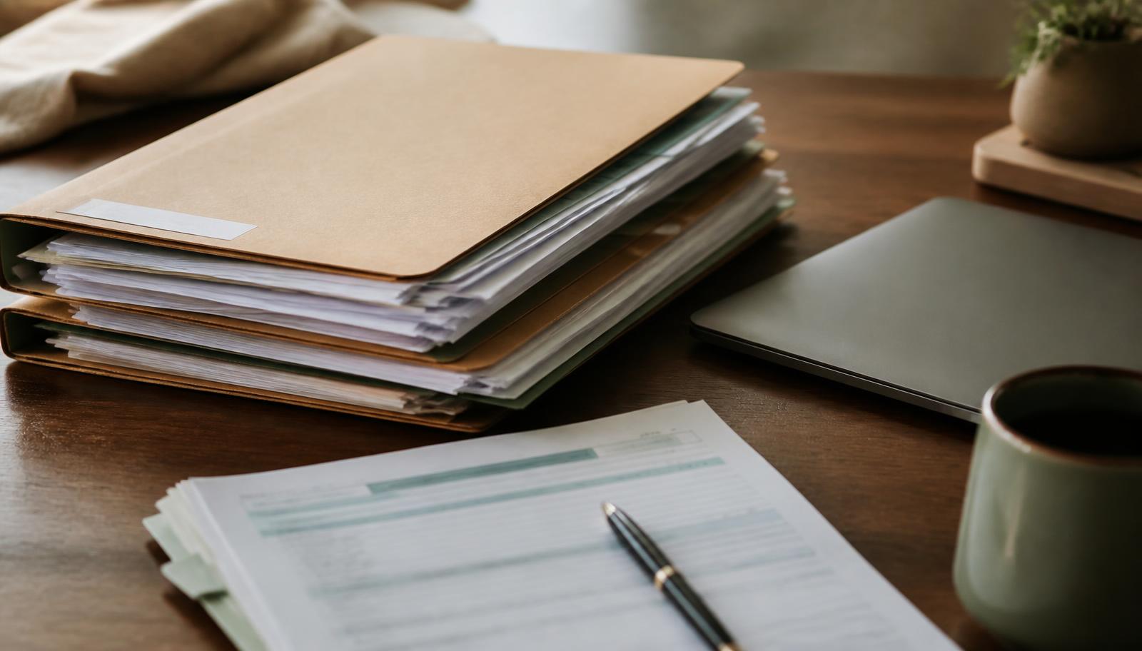 Old manila employment folders and financial paperwork stacked beside a closed laptop on a wooden desk